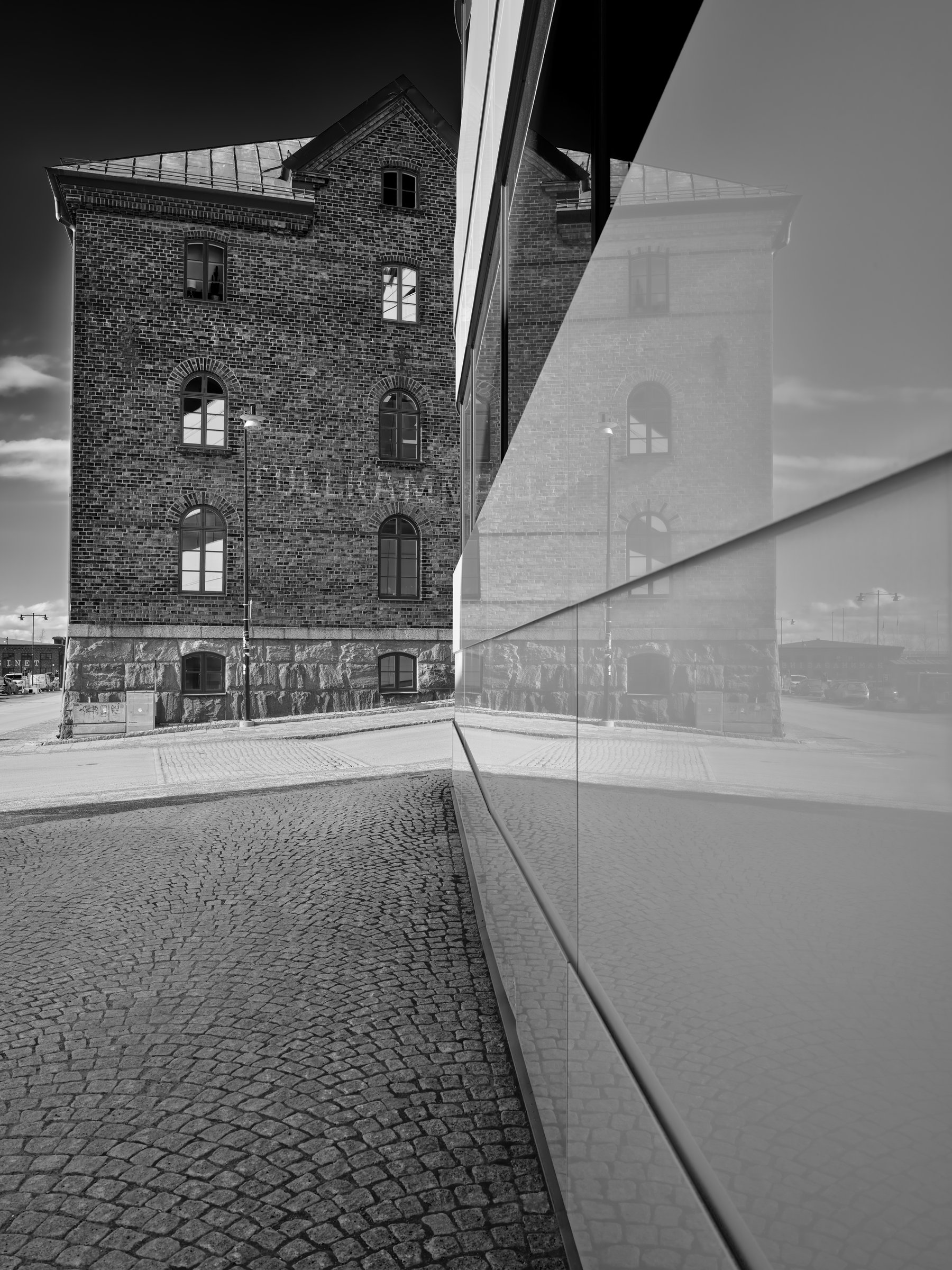 Svartvit bild av ett tegelhus som speglas i en glasfasad längs en stenlagd gata.