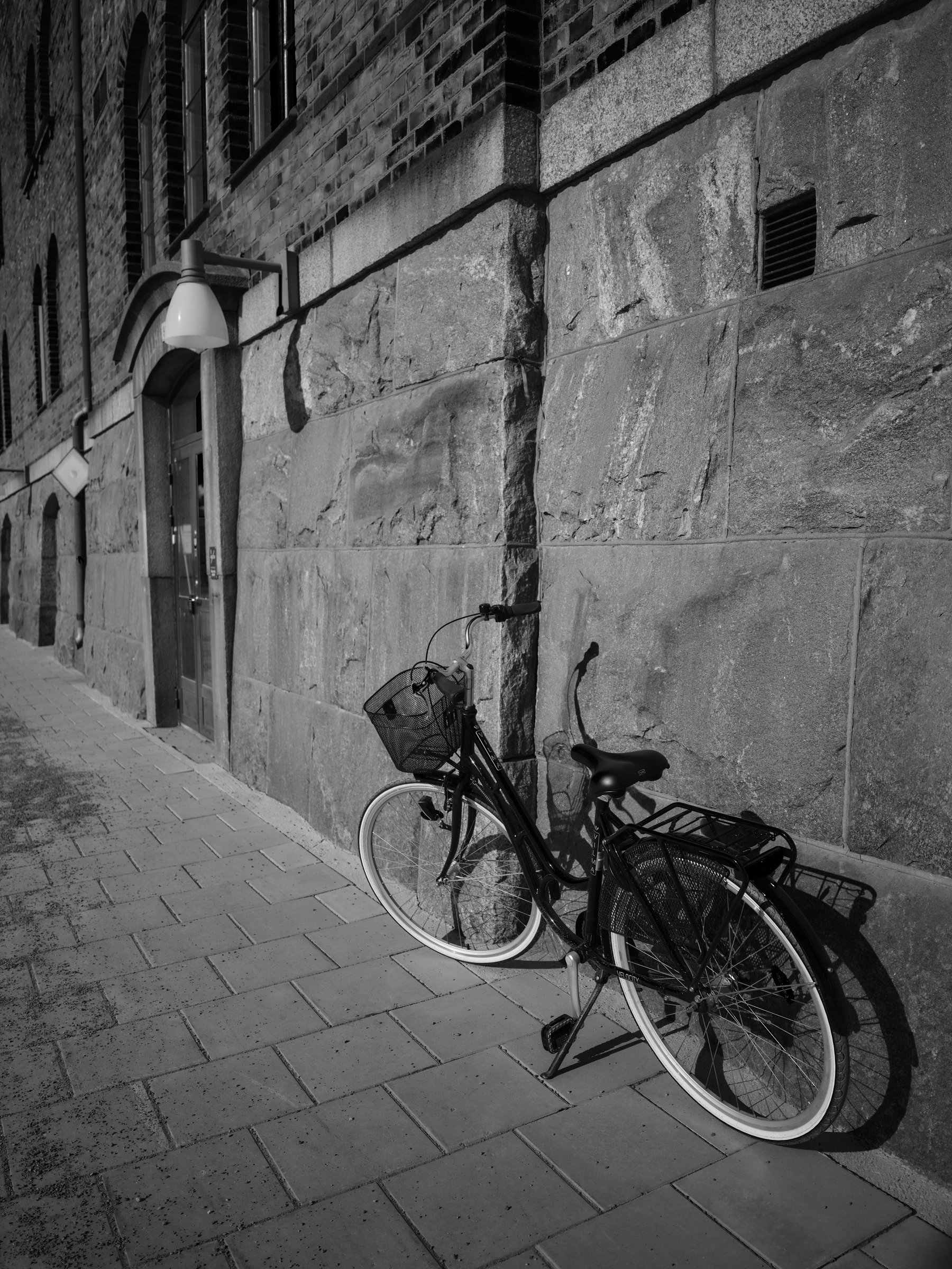 Svartvit bild av en cykel som står parkerad mot en grov stenvägg på en smal gata.