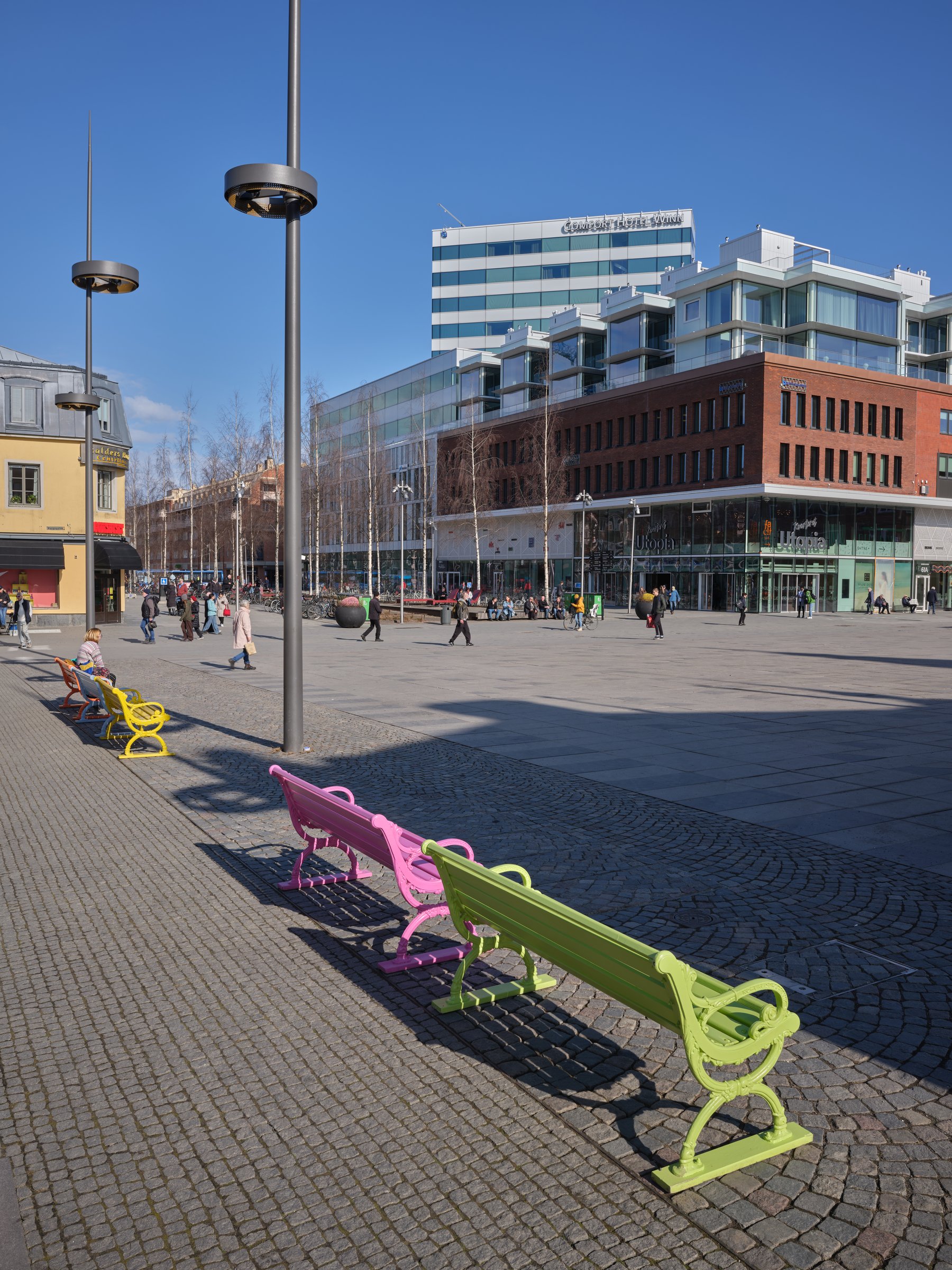 Torg i Umeå med färgglada bänkar i rad och moderna byggnader i bakgrunden, blå himmel. -hero